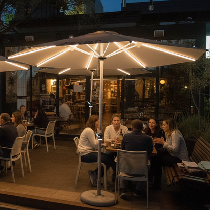 Outdoor patio dining area at dusk with two large illuminated patio umbrellas shading tables and seated people in front of a restaurant or bar.